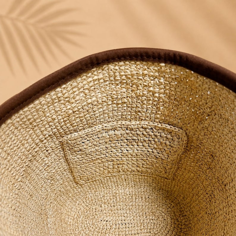 Pu&ograve; includere: Un cappello da pescatore in paglia intrecciata color beige naturale con un bordo in tessuto marrone scuro. L'interno del cappello presenta una toppa in paglia intrecciata coordinata cucita sul lato, che mette in risalto la trama del tessuto.