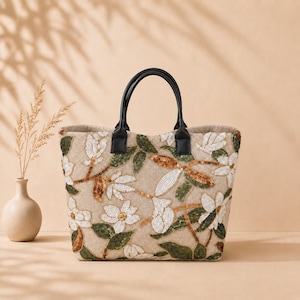 Puede incluir: Un bolso tote de color beige con un patrón floral bordado con lentejuelas blancas, verdes y bronce. El bolso cuenta con dos asas negras fijadas en el borde superior, lo que le proporciona una forma estructurada para llevar objetos.