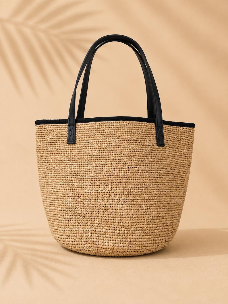 Pu&ograve; includere: Una borsa tote in paglia intrecciata con base arrotondata e una texture in fibra naturale color marrone chiaro. La borsa presenta due manici in similpelle nera fissati al bordo superiore, rifinito con un bordo in tessuto nero solido.