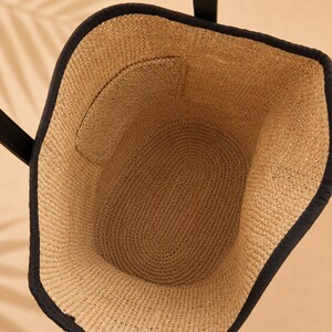 Pu&ograve; includere: Una vista dall'alto di una borsa tote in paglia intrecciata color beige naturale con un bordo in tessuto nero lungo il margine superiore. La borsa presenta due manici neri e una tasca interna integrata nella parete intrecciata della borsa.
