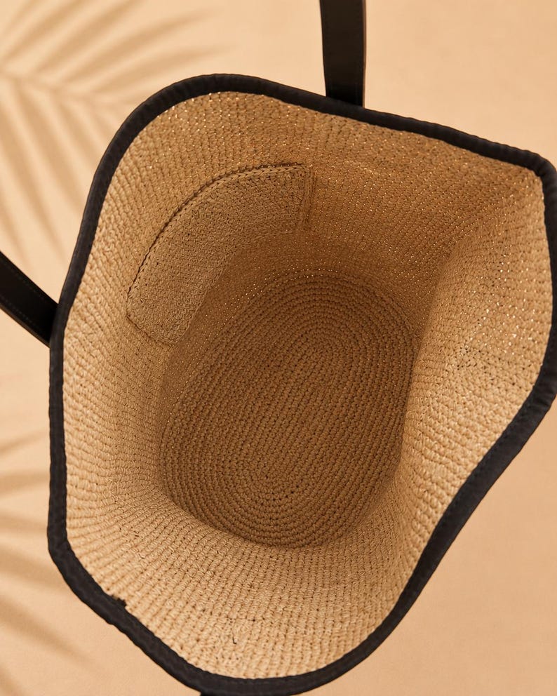 Pu&ograve; includere: Una vista dall'alto di una borsa tote in paglia intrecciata color beige naturale con un bordo in tessuto nero lungo il margine superiore. La borsa presenta due manici neri e una tasca interna integrata nella parete intrecciata della borsa.