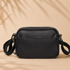 May include: A black rectangular crossbody bag with a textured surface, featuring a front slip pocket, a top zipper closure, and antique brass-toned metal hardware attachments for the adjustable shoulder strap on both sides of the bag.
