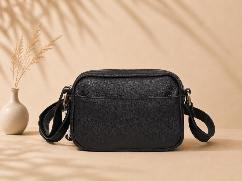 May include: A black rectangular crossbody bag with a textured surface, featuring a front slip pocket, a top zipper closure, and antique brass-toned metal hardware attachments for the adjustable shoulder strap on both sides of the bag.
