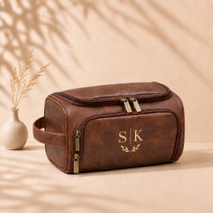 May include: A brown leather toiletry bag featuring a side handle, two zippered compartments, and gold-toned hardware. The front of the bag is personalized with the gold-colored letters S and K separated by a vertical line, with a small leaf motif below.