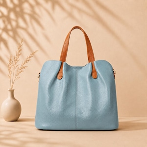 May include: A light blue tote bag with a textured surface, featuring two tan brown handles attached to the top. The bag has a vertical seam running down the center of the front panel and a structured base, providing a clean and functional design.