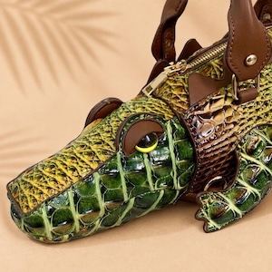 May include: A handbag shaped like a crocodile with a glossy green and yellow textured exterior. The bag features brown leather handles, a gold-tone metal zip closure, and a stylised eye detail with a yellow iris and black pupil.