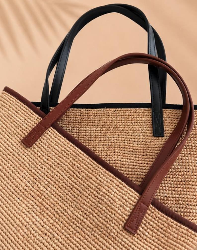 Pu&ograve; includere: Una borsa tote in paglia intrecciata con una texture color marrone chiaro naturale, rifinita con un bordo marrone scuro lungo il bordo superiore e due paia di manici. Una coppia di manici &egrave; nera, l'altra &egrave; marrone scuro, entrambe fissate.