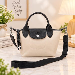 May include: A small beige canvas handbag featuring black leather handles and a black leather flap closure with a silver-tone snap button. The bag includes a detachable black fabric shoulder strap and a decorative black cord pull on the zipper.