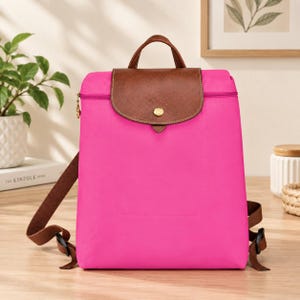 May include: A bright pink backpack featuring a brown textured leather flap with a gold-tone snap button closure. The bag includes a top handle, matching brown adjustable shoulder straps, and a horizontal zipper closure across the top section.