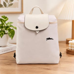 May include: A light beige backpack featuring a top handle and a fold-over flap with a snap button closure. The flap has a textured finish and a small embossed logo. A black embroidered horse logo is visible on the front of the main body.