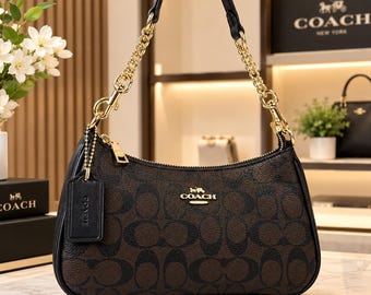 Puede incluir: Un bolso de mano Coach en marrón y negro con el patrón de monograma característico, una correa de cadena dorada, un asa de cuero negro y una etiqueta de cuero a juego. El bolso tiene cierre de cremallera y el logo de Coach en dorado.