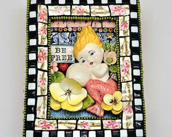 Könnte beinhalten: Ein Mosaik-Rahmenbild mit einer Meerjungfrau mit einem rosa Schwanz und gelben Haaren. Die Meerjungfrau hält eine Perle und hat einen süßen Gesichtsausdruck. Das Kunstwerk hat einen schwarz-weißen Schachbrett-Rand mit rosa Rosen und den Worten "BE FREE" in der Mitte.