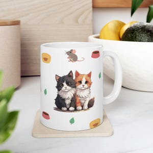 Pode incluir: Uma caneca de cerâmica branca com uma ilustração central de um gato fofo preto e branco sentado ao lado de um gato fofo laranja e branco. A caneca é decorada com pequenos ícones de um rato cinza, tigelas de comida rosa, camas amarelas e folhas verdes.