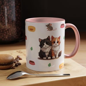 Pode incluir: Uma caneca de cerâmica branca com interior e alça cor-de-rosa, apresentando um padrão repetitivo de dois gatinhos felpudos, um pequeno rato castanho, camas amarelas para animais, taças cor-de-rosa e folhas verdes na superfície.