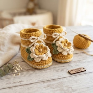 Könnte beinhalten: Ein Paar senfgelbe Häkelschuhe mit weißen Querstreifen, einer weißen Häkelblume mit einer zentralen Holzperle und grünen Blättern. Ein kleines Holzschild mit der Aufschrift HEARTMADE CROCHET liegt daneben.