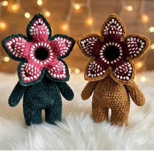 May include: Two plush crochet dolls featuring a five-petaled flower-shaped head with a dark centre, textured pink and white teeth-like accents, and a soft velvet-like yarn body. One doll is dark green, and the other is a light brown tan colour.
