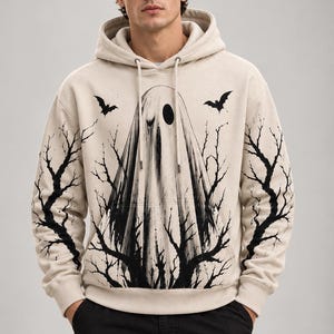 Könnte beinhalten: Ein beigefarbener Kapuzenpullover mit einem schwarzen Grafikdruck eines in Stoff gehüllten Geistes, umgeben von kahlen Baumzweigen und zwei fliegenden Fledermäusen. Der Hoodie verfügt über eine Kängurutasche, lange Ärmel und passende Kordelzüge.