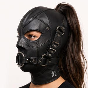 Puede incluir: Una capucha de cuero negro con diseño de cara completa y aberturas para ojos, nariz y boca. Incluye un bocado de anillo metálico, correas con hebillas ajustables en los laterales y herrajes plateados. Se aprecian las letras RL con una corona.