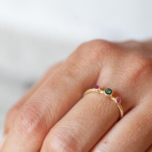 Pode incluir: Um anel de ouro com três pequenas pedras preciosas. As pedras preciosas são verdes e vermelhas.