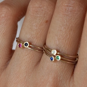 Pode incluir: Três anéis de ouro com pequenas pedras preciosas. Os anéis estão empilhados em um dedo. As pedras preciosas são vermelha, azul e verde.