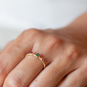 Pode incluir: Um anel de ouro com três pequenas pedras preciosas. O anel tem duas pedras vermelhas e uma pedra verde.