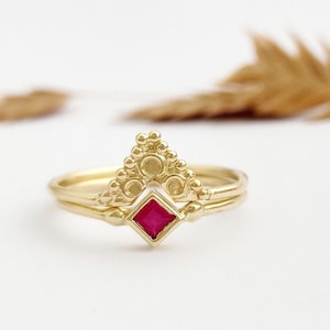Pode incluir: Um anel de ouro com uma gema vermelha engastada em forma quadrada. O anel tem um design delicado e intrincado com pequenos círculos.