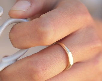 May include: A gold-colored ring with a flat top and sides. The ring is worn on a finger. A white teacup with a gold rim is held in the same hand.