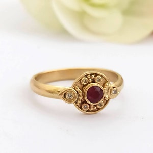 May include: A gold ring with a red gemstone surrounded by smaller white gemstones. The ring has a detailed design.