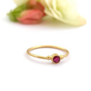 Puede incluir: Un delicado anillo de oro con una sola piedra preciosa roja redonda engastada en el centro.