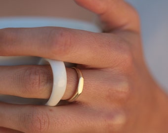 May include: A gold band ring with a slightly rounded, flat top. The ring is worn on the finger of a hand holding a white mug.