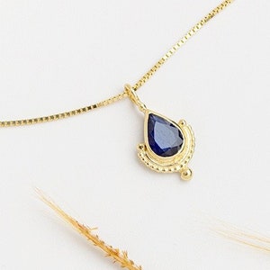 May include: A gold chain necklace with a teardrop-shaped pendant featuring a dark blue gemstone surrounded by a gold filigree design.