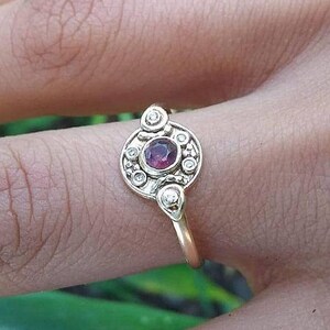 以下が含まれることがあります： 赤い宝石が、小さな白い宝石で囲まれた金の指輪。繊細で複雑なデザインの指輪です。