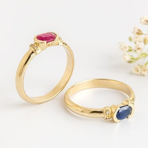 K&ouml;nnte beinhalten: Zwei goldene Ringe mit ovalen Edelsteinen. Der Ring links hat einen roten Rubin, und der Ring rechts hat einen blauen Saphir.