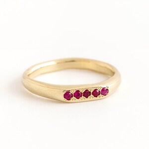 Può includere: Un anello a fascia d'oro con una fila di piccoli rubini rossi incastonati al centro.