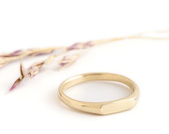 May include: A simple gold band ring with a flat, rectangular top. The ring is on a white background.