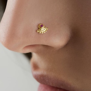 Może przedstawiać: Złoty kolczyk do nosa o wachlarzowatym kształcie z małym, okrągłym, czerwonym kamieniem szlachetnym pośrodku. Biżuteria jest noszona w lewym nozdrzu nosa osoby.