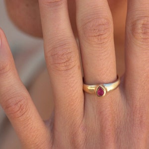 May include: A gold ring with a teardrop-shaped, red gemstone. The ring is on a person's finger, showcasing the jewelry's design and color. The ring's band is a simple, polished gold.