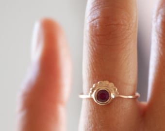 Op de afbeelding: Een gouden ring met een kleine, ronde, donkerrode edelsteen gezet in een delicaat, gegolfd ontwerp.