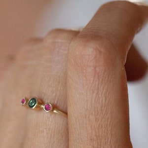 Puede incluir: Un anillo de oro con tres pequeñas piedras preciosas. El anillo tiene dos piedras rosas y una piedra verde.