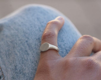 Könnte beinhalten: Ein silberner Siegelring mit einer glatten, ovalen Oberseite. Der Ring wird an einem Finger einer Hand getragen, die auf einer blauen Jeans liegt.