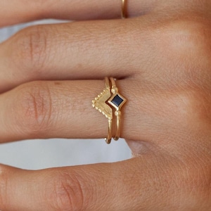 Op de afbeelding: Een gouden ring met een zwarte diamantvormige steen. De ring is ontworpen met een chevron-vorm en wordt aan een vinger gedragen.