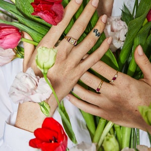 K&ouml;nnte beinhalten: Eine Hand tr&auml;gt drei goldene Ringe mit Rubin- und Diamantbesatz. Die Hand h&auml;lt einen Strau&szlig; roter und wei&szlig;er Blumen.