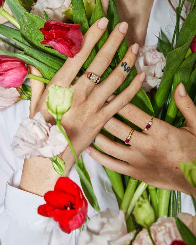 K&ouml;nnte beinhalten: Eine Hand tr&auml;gt drei goldene Ringe mit Rubin- und Diamantbesatz. Die Hand h&auml;lt einen Strau&szlig; roter und wei&szlig;er Blumen.