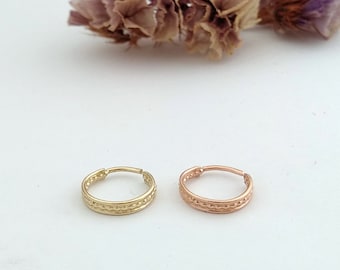 May include: Two small, decorative hoop earrings. One is gold-colored, the other is rose gold. Both have a textured, repeating pattern around the outer edge. The earrings are displayed on a white surface with dried flowers in the background.