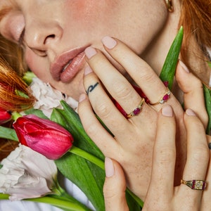 K&ouml;nnte beinhalten: Eine Frauenhand tr&auml;gt drei goldene Ringe mit roten Edelsteinen. Die Ringe befinden sich an ihrem Zeige-, Mittel- und Ringfinger. Die Hand ruht auf einem Strau&szlig; rosa und roter Tulpen.