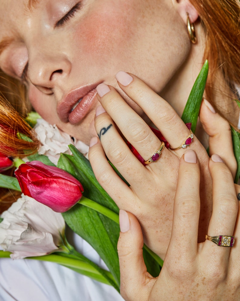 K&ouml;nnte beinhalten: Eine Frauenhand tr&auml;gt drei goldene Ringe mit roten Edelsteinen. Die Ringe befinden sich an ihrem Zeige-, Mittel- und Ringfinger. Die Hand ruht auf einem Strau&szlig; rosa und roter Tulpen.
