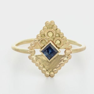 May include: A gold ring featuring a blue gemstone set within a geometric design. The ring is embellished with small beads and circles.
