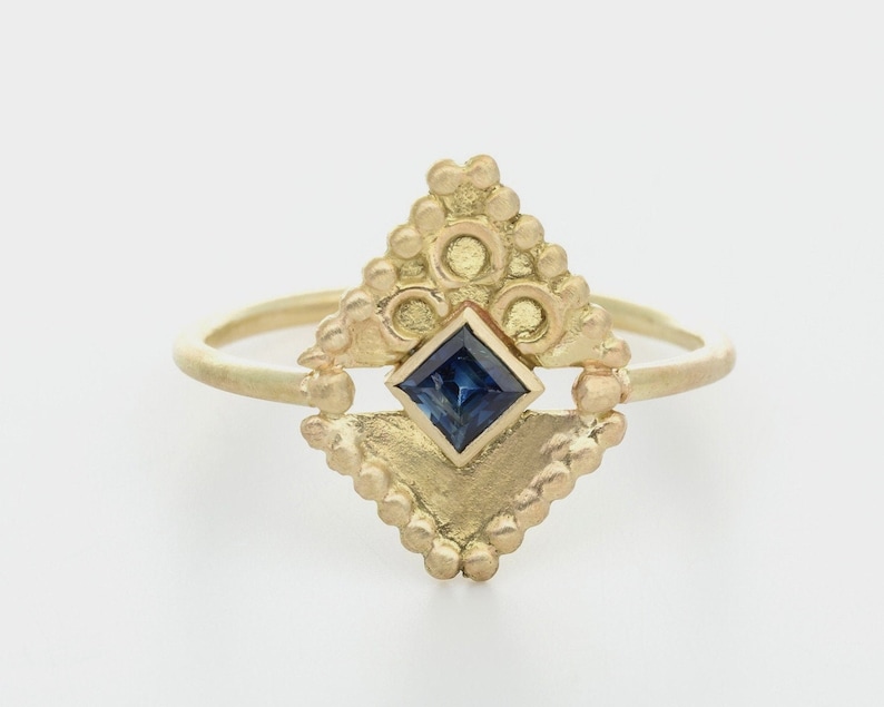 May include: A gold ring featuring a blue gemstone set within a geometric design. The ring is embellished with small beads and circles.