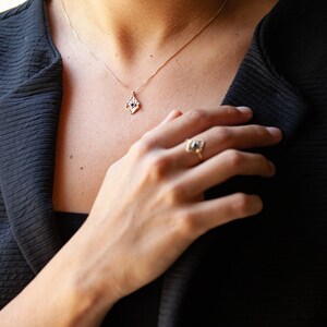 May include: A gold necklace with a small, teardrop-shaped pendant featuring a blue gemstone. The pendant is set in a delicate, geometric design. The necklace is worn by a person with a ring on their finger, also featuring a blue gemstone.