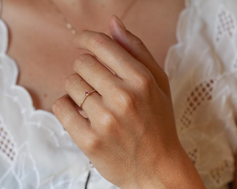 May include: A delicate gold band ring with a small, bright red gemstone set in the centre.